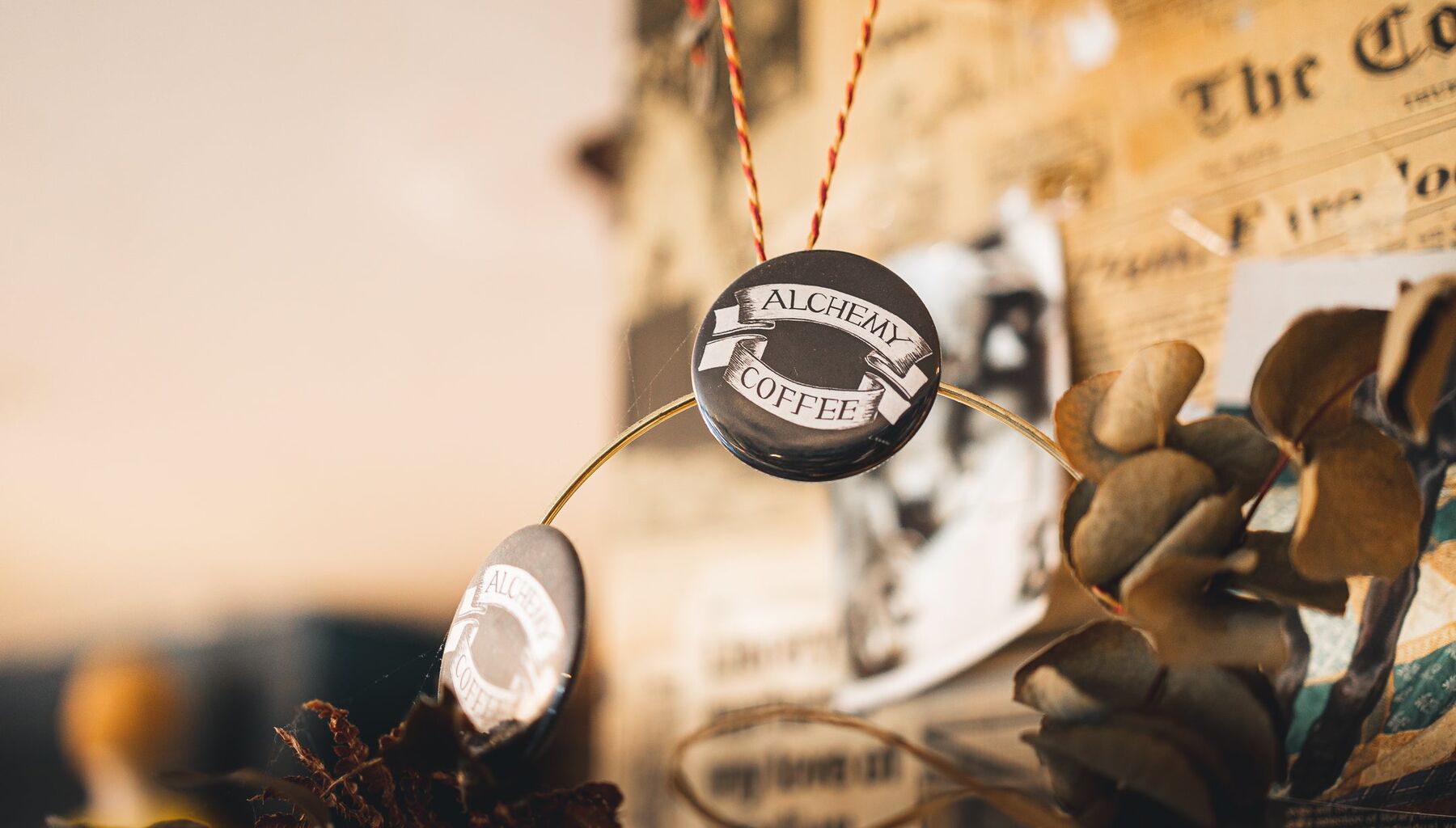 Two Alchemy Coffee pin badges hung on a ring in front of old Cork Examiner pages and dried eucalyptus